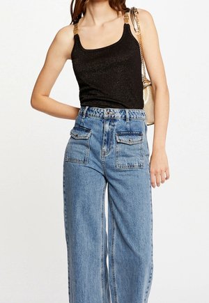 Kobieta ubrana w czarny top bez rękawów z złotymi detalami na ramiączkach, niebieskie dżinsy z wysokim stanem i szerokimi nogawkami oraz dużymi przednimi kieszeniami, nosząca złotą torebkę na ramię.