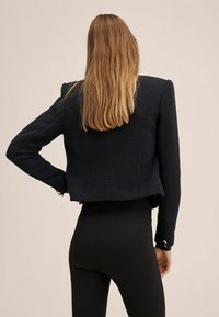 Marineblau cropped Jacke aus strukturiertem Stoff, mit betonten Schultern und goldfarbenen Hardwareelementen an den Ärmeln; kombiniert mit schwarzen Leggings.