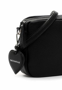 Melnas teksturētas ādas plecu soma ar rāvējslēdzēju, piesaistītu sirds formas piekariņu ar zīmolu "EMILY & NOAH" uz balta fona.