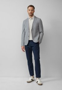 Leichtgrauer Blazer für Herren mit zwei Knöpfen, kombiniert mit einem weißen Polohemd und marineblauen Chinos. Weiße und braune Sneaker runden das Outfit ab.