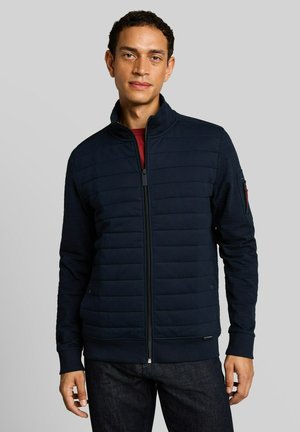 Mann trägt eine dunkelblaue gesteppte Jacke mit Reißverschluss und hohem Kragen sowie dunkle Jeans und steht vor einem schlichten grauen Hintergrund.