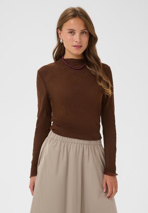 Jeune femme aux longs cheveux châtain clair porte un haut marron à manches longues, une jupe beige taille haute, un collier de perles violet et des boucles d'oreilles en perles.