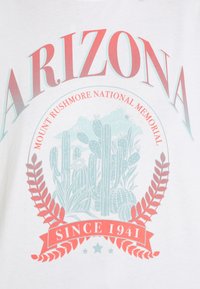 Bílý tričko s nápisem "ARIZONA" v červené a tyrkysové barvě, se grafikou kaktusu a nápisy "MOUNT RUSHMORE NATIONAL MEMORIAL" a "SINCE 1941" v dekorativním písmu.