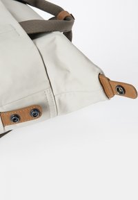 Lichtbeige tas met een gestructureerde stof, met bruin leren accenten en metalen drukknoopsluitingen. Het ontwerp heeft verstelbare banden en een zijnaad.