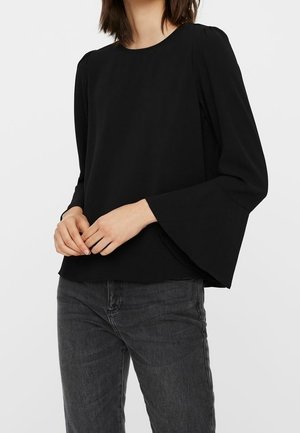 Nainen pukeutunut mustaan pitkähihainen puseroon, jossa on leveät kellohihat, ja tummanharmaisiin farkkuihin, seisoo käsien rentoina puristettuina.