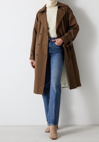 Bruine trenchcoat, crèmekleurige gebreide trui, high-waisted blauwe jeans en beige hakken. Heeft dubbel-breasted knopen en een relaxte pasvorm.
