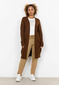Brown knitted long cardigan with open front and pockets, paired with beige sweatpants and a white graphic t-shirt. White sneakers complete the look.