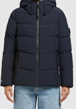 Veste matelassée puff bleu marine avec capuche, fermeture éclair avant, poches latérales zippées et cordons de serrage ajustables, portée avec un jean bleu clair.