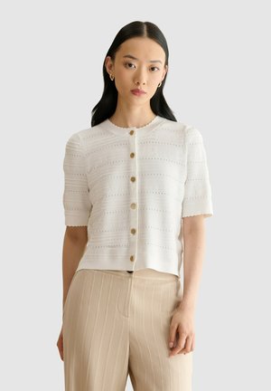 Vrouw met lang donker haar draagt een wit kort mouwen blouse met knopen en beige hoge taille broek met verticale strepen, staand voor een effen achtergrond.