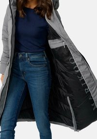 Szara kurtka puchowa z kapturem, z przednim zamkiem błyskawicznym, czarną podszewką i kieszeniami bocznymi; noszona na ciemnoniebieskiej koszuli i dżinsach.