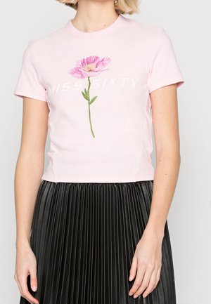 Kobieta ubrana w jasnoróżowy krótki t-shirt z dużym różowym kwiatem i napisem "MISS SIXTY", w połączeniu z czarną plisowaną spódnicą.