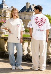 Giovane donna e uomo in piedi all’aperto che indossano t-shirt bianche con grafiche di cuori, farfalle e testo, vicino a una balaustra in pietra storica.