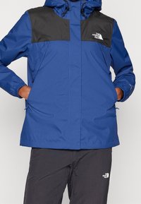 Veste imperméable bleue avec panneaux d'épaule noirs, capuche avec cordon de serrage, fermeture éclair à l'avant et poches latérales. Tissu épais et texturé pour assurer la durabilité.