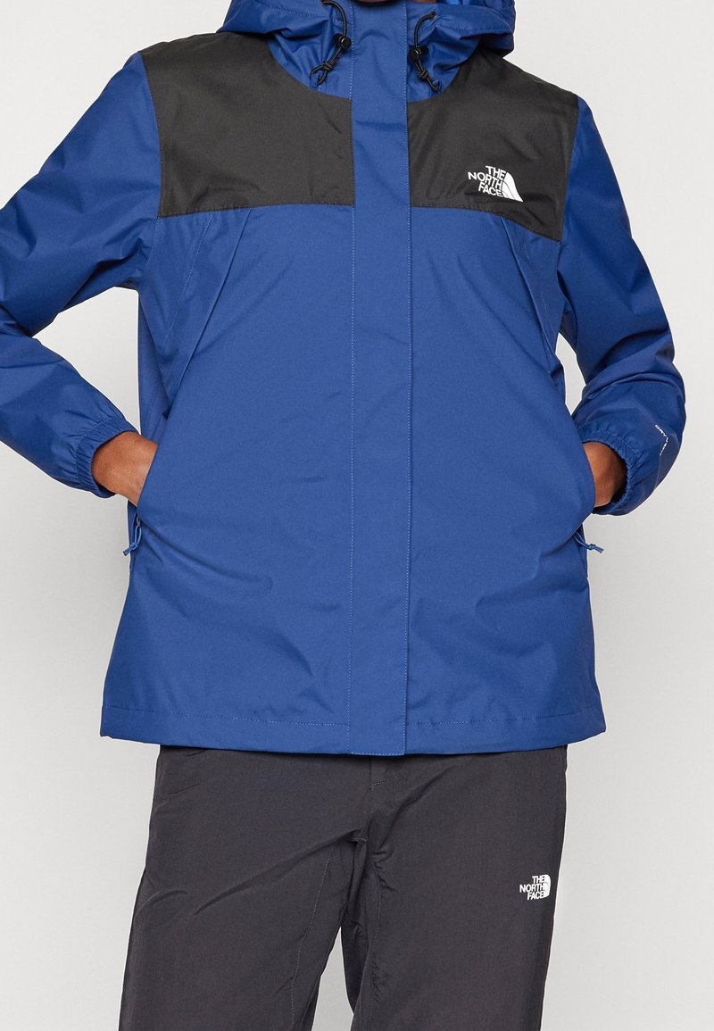 Veste imperméable bleue avec panneaux d'épaule noirs, capuche avec cordon de serrage, fermeture éclair à l'avant et poches latérales. Tissu épais et texturé pour assurer la durabilité.