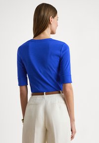 Krátky vrchný diel v kráľovskej modrej farbe s trojštvrťovými rukávmi, okrúhlym výstrihom a hladkou textúrou, skombinovaný s béžovými nohavicami s vysokým pásom a hnedým opaskom.