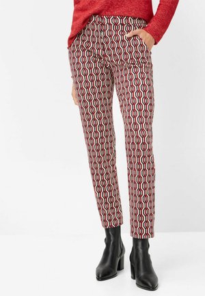 Nainen, jolla on päällään kuvioidut, kapealahkeiset housut, joissa on punainen, musta ja valkoinen geometrinen kuvio, mustat nilkkurit ja punainen neule, seisoo käsi taskussa.