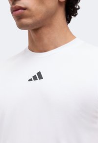 Man draagt een wit shirt met ronde hals en een klein zwart drie-strepen logo op de bovenborst tegen een effen achtergrond.
