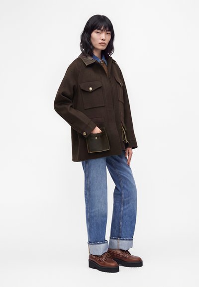 Femme portant une veste oversize marron foncé avec des poches, un jean bleu avec revers et des chaussures compensées marron, debout devant un fond blanc uni.