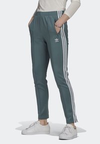 Pantaloni sportivi teal con logo Adidas bianco, tasche laterali con zip e tre strisce bianche lungo le gambe esterne. Design aderente e affusolato.