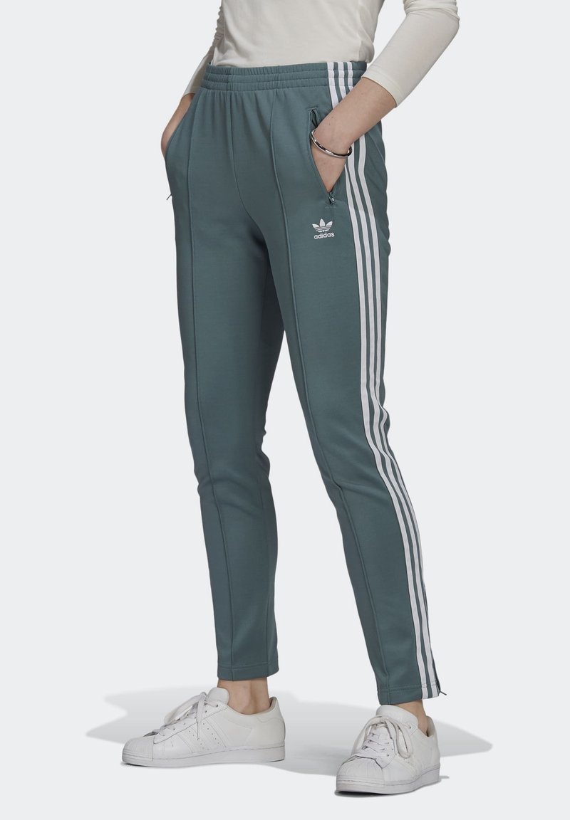 Pantaloni sportivi teal con logo Adidas bianco, tasche laterali con zip e tre strisce bianche lungo le gambe esterne. Design aderente e affusolato.