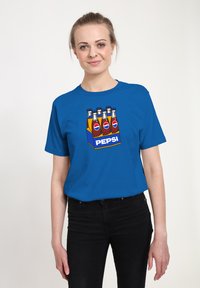 Žena v modrém tričku s grafikou šesti lahví Pepsi v žlutém nosiči a černých kalhotách na jednoduchém pozadí.