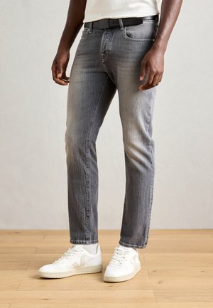 Grijze spijkerbroek met een slim fit, met een vervaagde afwerking. Gepaard met witte sneakers, tegen een achtergrond van een houten vloer.