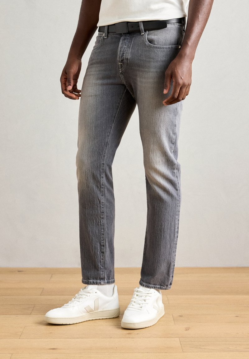 Jeans din denim gri cu un croi slim, având un finisaj decolorat. Combinat cu teniși albi, pe un fundal de podea din lemn.