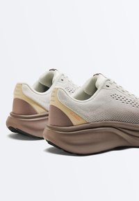 Para beżowo-brązowych sneakersów z poduszkami, o teksturowanych dzianinowych cholewkach, grubych podeszwach i napisem "OVER835" blisko tylnych części butów.