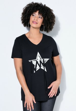 Femme aux cheveux bouclés portant de grandes boucles d'oreilles anneaux et un t-shirt noir à col en V avec une étoile blanche décorée d'effets usés et des cordons noirs.