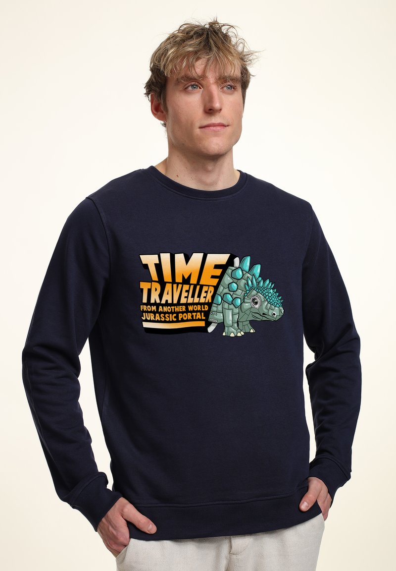Felpa blu navy con un design grafico che presenta un dinosauro in stile cartone animato e il testo "VIAGGIATORE DEL TEMPO DA UN ALTRO MONDO PORTALE GIURASSICO."