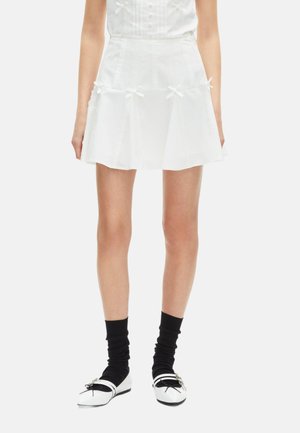 Jupe blanche évasée avec des détails en dentelle et de petits nœuds, fabriquée en matériau léger. Associée à des chaussettes noires et des chaussures noir et blanc.