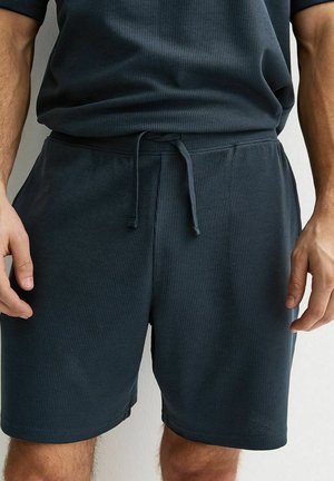 Męskie granatowe teksturowane szorty ze ściągaczem i pasująca koszula, widoczne od talii do połowy uda, z rękami swobodnie opuszczonymi wzdłuż ciała.