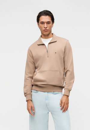 Junger Mann mit dunklen Haaren, der einen beigen Sweatshirt mit Viertelreißverschluss und hellblaue Jeans trägt, steht vor einem schlichten grauen Hintergrund.