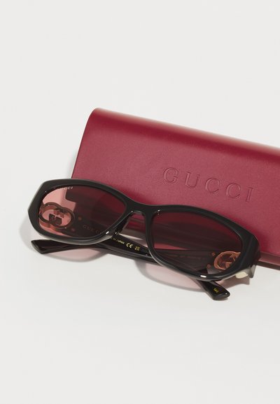 Slnečné okuliare Gucci s čiernymi rámami a gradientnými ružovými sklami, ležiace na tmavočervenom puzdre s vyrazeným logom.