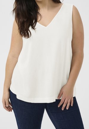 Frau trägt ein ärmelloses weißes V-Ausschnitt-Top und dunkelblaue Jeans, steht mit einer Hand auf dem Oberschenkel.