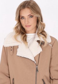 Braune Kunstlederjacke mit einem plüschigen weißen Schaffellkragen, Reißverschluss und Seitentaschen. Verfügt über eine entspannte Passform und saubere Nähte.