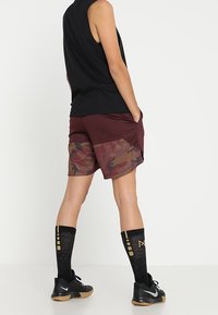 Svart ärmlös topp, vinröda och kamouflagebyxor, svarta strumpor med guldaccenter, svarta sneakers, sedd från baksidan.