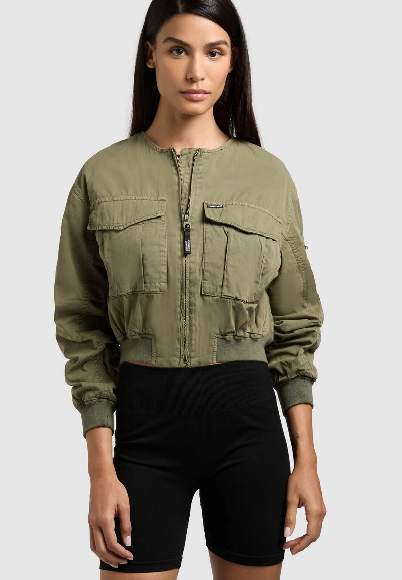 Femme aux longs cheveux bruns portant une veste zippée vert olive avec de grandes poches frontales et un short noir moulant sur un fond uni.