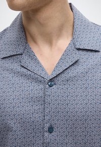 Lühikeste varrukatega särk, millel on sinise ja valge geomeetriline muster, notsisäärisega krae ja üks sinine nööp lainel.