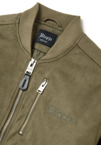 Chaqueta bomber de color verde oliva hecha de suave ante, con un cuello acanalado, cremalleras plateadas y el término "Utopía" bordado en cursiva.