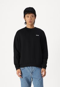 Svart genser med rund hals, lange ermer og ribbestrikket kant. Hva som kjennetegnes av en liten hvit logo på venstre bryst. Kombinert med lyseblå jeans.
