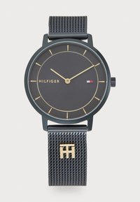 Montre avec bracelet en maille noire et accents dorés. Le cadran rond noir présente des index et des aiguilles dorées. Le nom de la marque "HILFIGER" est affiché.