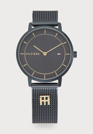 Black mesh strap watch with gold accents. Round black dial features gold hour markers and hands. Brand name "HILFIGER" displayed.