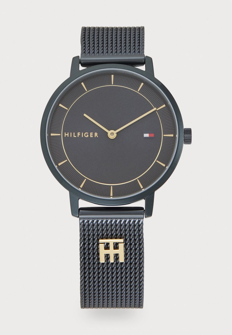 Fekete hálós pántos óra arany díszítéssel. Kerek fekete számlap arany órajelzőkkel és mutatókkal. A márka neve: "HILFIGER".