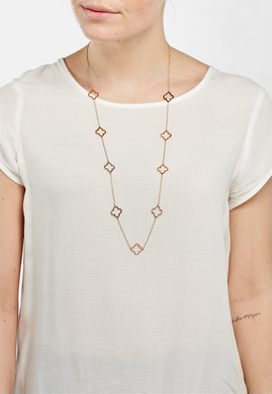 Collana lunga in tono dorato con pendenti a forma di fiore alternati, con ritagli e centri solidi, indossata su una maglietta bianca.