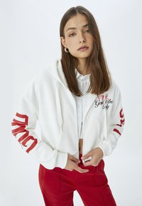Sudadera blanca con cremallera y texto gráfico rojo en las mangas. Llevada sobre una camiseta blanca, combinada con pantalones rojos de cintura alta. Textura de tela suave.