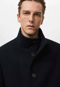 Granatowy płaszcz z wełny z wysokim kołnierzem, zapinany na guziki, wykonany z fakturowanej tkaniny. Posiada trzy czarne guziki i czysty, minimalistyczny design.