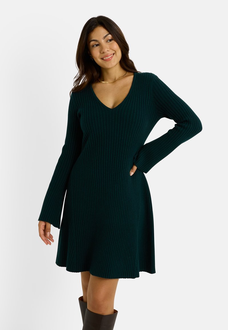 Robe en tricot côtelé vert profond avec un décolleté en V, des manches longues et une jupe évasée. Le matériau semble doux avec une finition texturée.