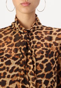 Brūns un smilšu krāsas leopardu raksta blūze ar sasietu kakla daļu, kas izgatavota no viegla, caurspīdīga auduma un mīkstas tekstūras.