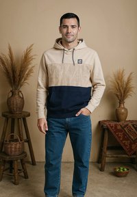 Hoodie met beige bovenkant, marineblauwe onderkant en een bladpatroon; gecombineerd met een blauwe spijkerbroek. Model staat met handen in de zakken, neutrale achtergrond.